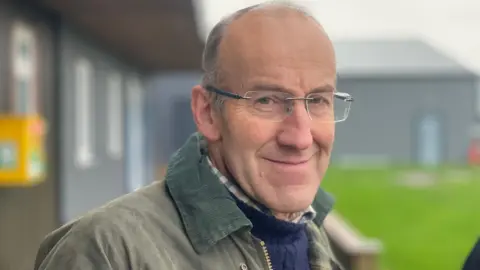 A man wearing glasses and a green waxed cotton coat is looking into camera. There are buildings in the background