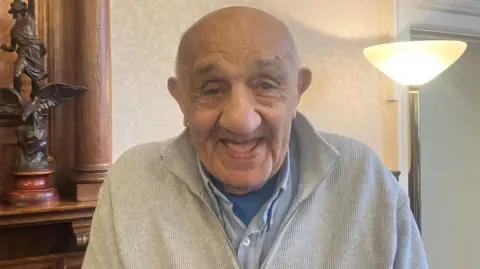 Rugby league legend Sir Billy Boston who is wearing a grey ribbed zip up jumper with a blue shirt and t-shirt underneath at a civil ceremony in his honour. He is smiling.