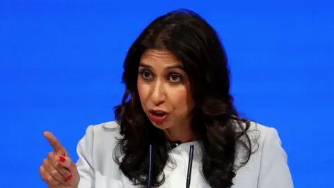 Reuters Suella Braverman sitting in front of a plain blue background, pointing with her right index finger.