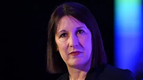 Getty Images Rachel Reeves. She has black hair, brown eyes and red lipstick. She wears a balck suit jacket. A purple glow highlights one half of her face. 