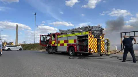Will Jefford/BBC A fire engine at the scene of a fire. Fire crews pull long water hose pipes. Thick smoke billows into the air behind them. Another fire engine is parked in the distance along with a silver car. 