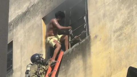 Man wearing just a pair of shorts climbs out of window and steps on ladder