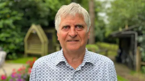 Rob Prow looking at the camera in a garden. He's wearing a patterned shirt, and has grey hair.