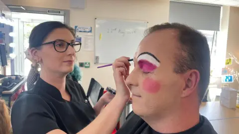 A man has pink eye make up applied by a make up artist wearing a black uniform. She has tied back brown hair and black glasses. 