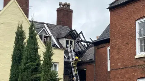A black and white timbered building seen from behind two other buildings, with part of its roof missing and timbers exposed to the sky