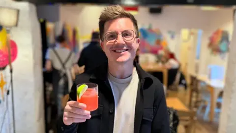 BBC / Josh Gorroño Chapman Henry Beercock looks into the lens smiling. He has dark brown hair and wears thin-rimmed glasses, a cream t-shirt and a dark overshirt. He is holding a bright pink cocktail garnished with a lime wedge, an Alcohull Free.