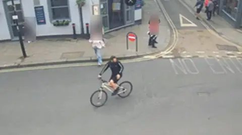 North Yorkshire Police A street scene captured by CCTV shows a cyclist riding towards the camera. On the corner of the street is a pub and pedestrians are blurred from view.