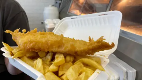Andrew Crook handout Close up of someone holding a portion of fish and chips in a cardboard take away box behind the counter of a fish and chip shop.