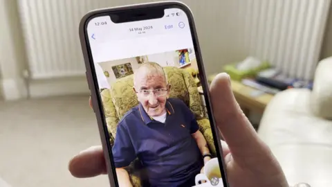 Supplied A close up of a photo of Mick Griffiths on a phone. He is sitting in a green armchair and smiling at the camera. He has a blue polo shirt on. 