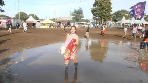 Amber Richards Amber stands in an orange-red dress and wellies, in a large puddle in the middle of a festival site