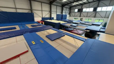 A warehouse which contains a foam pit and nine trampolines. There is padding on the floor all the way around the trampolines and there are thin mats on each of the trampolines. The far wall of the building has ground level and upper level windows, and there are roofs lights.