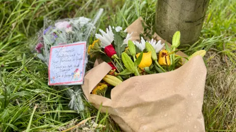 A bouquet of flowers have been placed on the grass. They are wrapped in brown paper. A small note has also been left which reads 'our deepest condolences to your families and friends. Thank you for your service'.