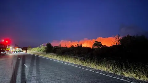 New Forest Police Fire in New Forest with orange glow seen to the right of a road and many emergency services in the distance.