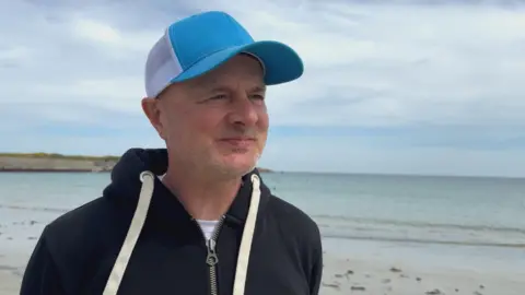 Alan Jenner standing on a beach. Waves are lapping behind him. Mr Jenner is wearing a black zip up hoodie and a blue cap.
