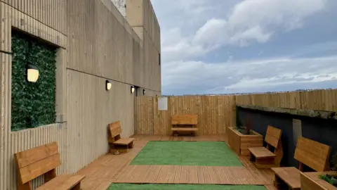 Wooden benches arranged around some decking and artificial turf with blue sky and white clouds behind.