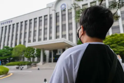 A woman with short hair stands facing a court building, her face is turned away from the camera