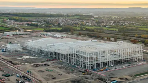 An aerial shot at sunset of the gigafactory steel frame on its construction site. There are houses and countryside in the distance. 