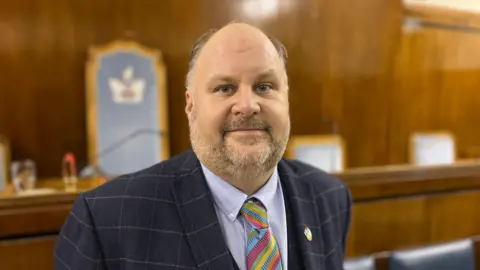 BBC Councillor Jim Robbins looks into the camera, smiling slightly. He is wearing a blue suit, shirt and tie - the tie is colourful with stripes.