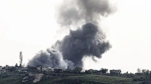 A large grey bundle of smoke rising from a village on a hillside. The smoke cloud fills most of the picture and is dramatic against the backdrop 