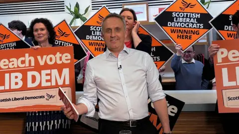 Alex Cole-Hamilton, standing in front of a group of LibDem supporters holding orange signs, smiling at the camera 