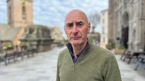 Deputy Steve Williams - A man with a bald head looking sternly at the camera. He is wearing a green quarter zip, with a blue spotted white shirt. 