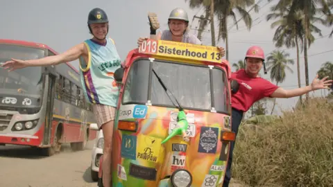 Three smiling women are in a colourful tuk-tuk on the side of a road. Two are hanging out the side with arms extended in a celebratory pose.