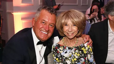 Getty Images Paul Burrell, wearing a black suit jacket, white shirt and a black bow tie, poses with his arm around Helen Worth. She has short blond hair and is wearing a floral dress. Both are smiling. 