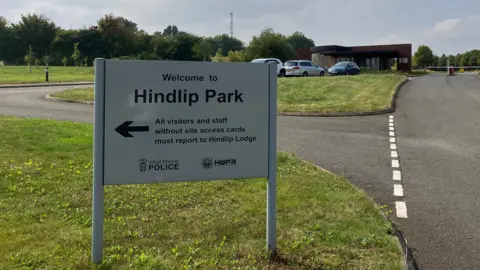 A sign at Hindlip Park, which is the headquarters of West Mercia Police, points visits and staff to the entrance. It is it a road junction surrounded by areas of grass. There are road markings and buildings and cars in the background.