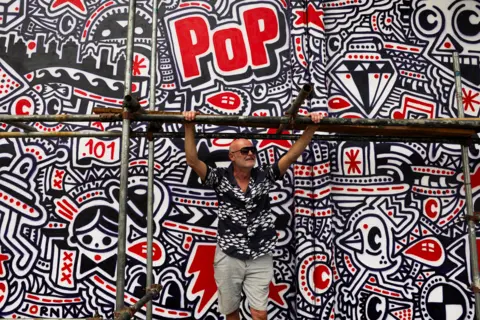 Sodiq Adelakun / REUTERS A man in a black and white shirt leans on scaffolding in front of a black, white and red abstract mural, featuring the word Pop in red.