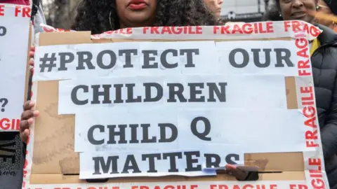 A protester holds a cardboard sign reading “#Protect our children, Child Q matters,” bordered with red “fragile” tape.