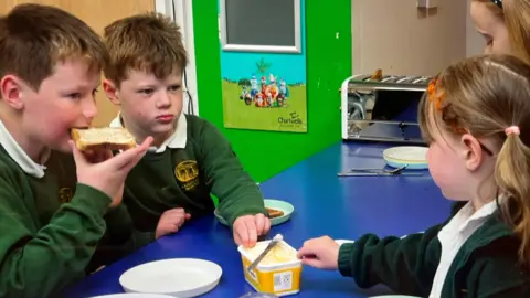Pupils wearing green school uniform eating breakfast seated at a blue table in a classroom setting. Two boys are on the left and one is eating toast, two girls are on the right. They are passing the butter to each other across the table, and a toaster is in the background.