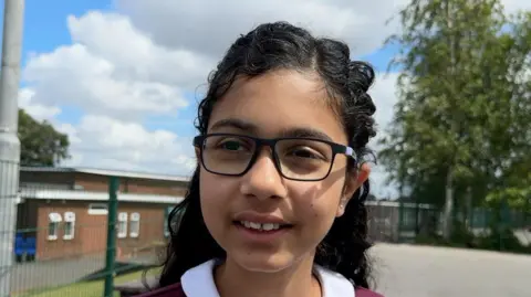 A close-up of a child's face. She is wearing glasses and appears to be in a school playground.