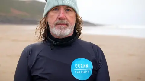 Phil Owen is wearing a wet suit with a blue logo reading ocean therapy. He has a grey beard and long light brown hair, under a light blue/green hat, which also says Ocean Therapy 