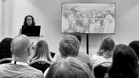 University of Bath A black and white picture of Dr Williamson standing at a podium in front of a seated audience, giving a presentation on moral injury. The screen beside her shows a soldier in a camouflage uniform and helmet, looking over a crowd of children and civilians.