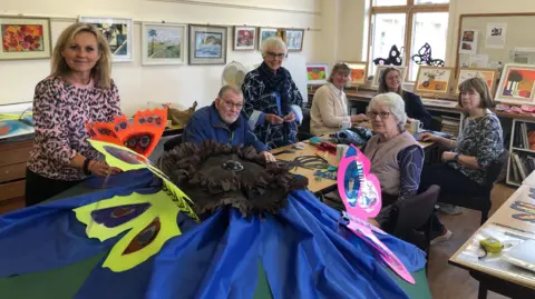 The group of members are seated around a table with one of the pieces on butterfly artwork on the table 