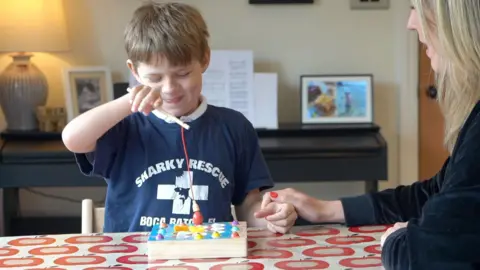 A boy wearing a blue 'Shark Rescue' t-shirt holds a toy fishing rod in his right hand, while "fishing" for magnetic toys on a table.