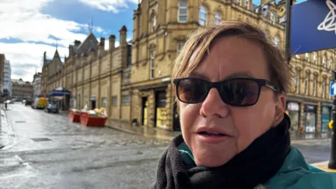 Gemma Dillon/BBC Image shows a lady wearing sunglasses and a scarf.
She is standing on a street in Halifax with the market hall behind her. There are skips on the road which are full of building waste. 