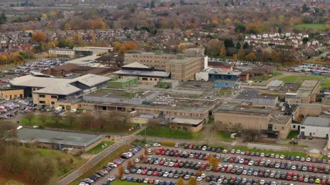 An aerial view of an area with a number of large brown buildings behind a large car park, with a scattering of trees in the distance