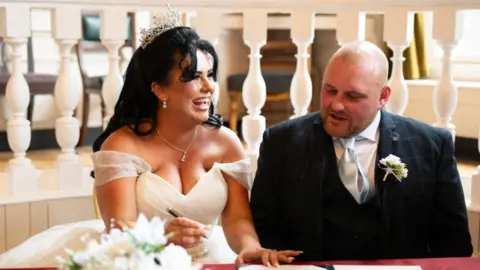 Mark Thompson @ MST Photography Shania Stanton and her husband on their wedding day signing the marriage schedule. She is on the left wearing a white dress with exposed shoulders and a tiara. She has long dark hair and is laughing and holding the pen. He is wearing a suit with a blue silk tie and a flower brooch. He is bald. There is a bouquet in the foreground. 