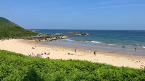 It shows a white sand beach with people on the sand and a few in the water. There are rocks in the background to the left.