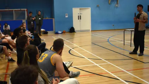 Suffolk New College On the right of the picture, Matt Hayden stands in the centre of the sports hall holding a microphone. A large number of students and staff are seated in front of him, listening.