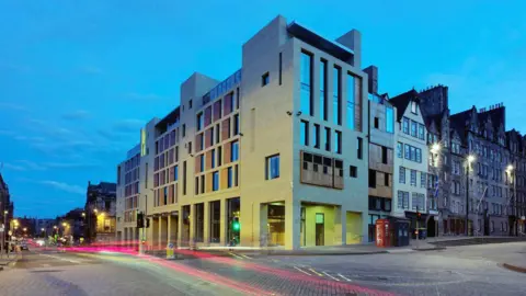 Radisson Hotels A modern sandstone building with large vertical windows on a cobblestone street in an urban setting at dusk, with light trails from passing vehicles and historic buildings in the background
