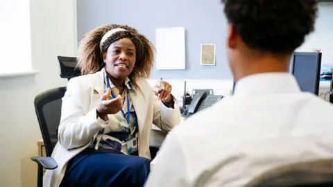 Getty Images A stock image shows a head teacher and pupil in an office setting, engaged in conversation. The head teacher is facing the camera, wearing a light-coloured blazer and patterned blouse, gesturing with her hands. The pupil is seen from behind, wearing a white shirt. A desk with office equipment, including a telephone and papers, is visible in the background.