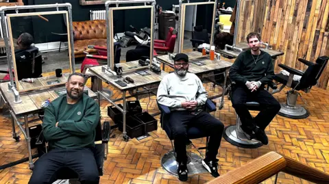 Three men sit in leather chairs in a barbershop. They have all spun around to look at the camera.