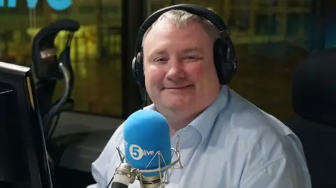 A man with white hair smiles at the camera. He is wearing black headphone on his head and a blue shirt. A blue microphone which says '5 live' sits in front of his chin. There is a clear window behind him, it's dark outside. 