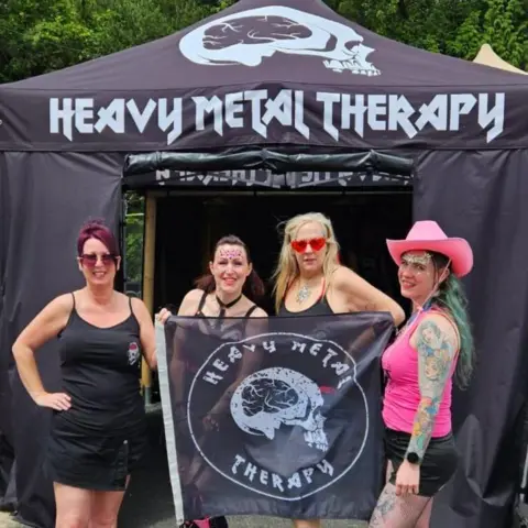 Heavy Metal Therapy Four women stand in front of a black tent branded Heavy Metal Therapy in white letters. They are also holding a flag with the organisation's name and logo - a white skull with a black brain - on it. 