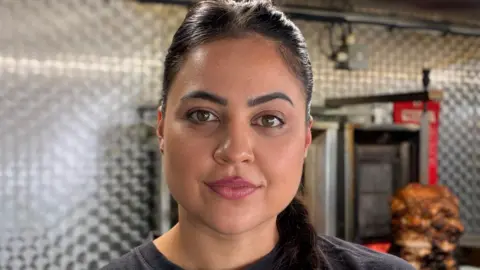 Sahar with tied-back black hair looks at the camera while inside her food van. There is an oven and a spit with roasted meat behind her. 