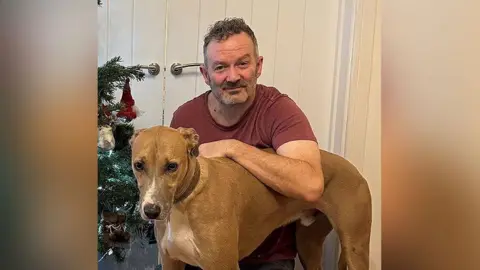 Alan Eastham, wearing a burgundy t-shirt and grey trousers, kneels down with his arm resting on his tan-and-white lurcher Alvin. They are in front of a set of white doors and a Christmas tree to their right. 