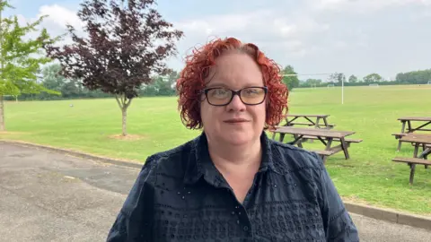 A woman standing by a playing field in a black top with red curly hair. There is a tree to the left of her head and picnic benches on the right.