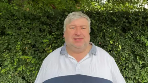 Contributed A man with grey hair and blue eyes smiling, wearing a white and navy blue polo shirt, standing in front of a leafy and dense green hedge.